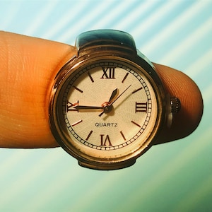 May include: A gold-toned ring watch with Roman numeral markings on a white face. The watch face reads "QUARTZ".