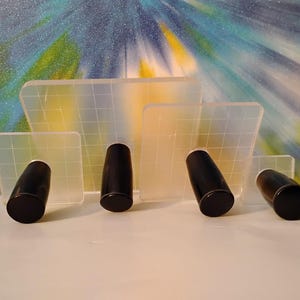 May include: A collection of clear, rectangular acrylic stamps with a grid pattern, paired with black, cylindrical handles. The stamps vary in size and are arranged in front of a colorful, abstract background.