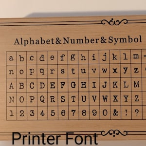May include: A wooden stamp with the text "Alphabet & Number & Symbol" and "Printer Font". The stamp features a grid of lowercase and uppercase letters, numbers, and symbols in a typewriter font. The stamp has a decorative border.