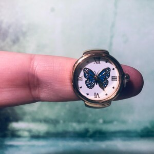 May include: A gold ring with a white face watch featuring a blue butterfly design. The watch face has Roman numeral hour markers and a gold-colored band.