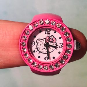 May include: Pink Hello Kitty watch with a rhinestone bezel and a white face. The watch has black hands and a Hello Kitty character in the center.