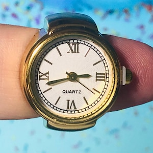 May include: A gold-toned ring watch with a white face and black Roman numeral hour markers. The watch face also has a black 'QUARTZ' label.