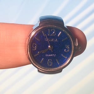 May include: A silver ring watch with a blue face and white numbers. The watch face has a sparkly blue background and the text "GOMIA QUARTZ" printed on it.