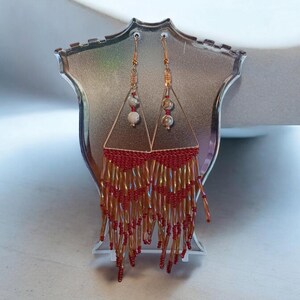 May include: A pair of gold dangle earrings with red and gold beads. The earrings feature a triangle shape with a small white and gray stone at the top.