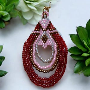 May include: A pair of long, beaded earrings with a teardrop shape. The earrings are made with red, pink, and gold beads. The earrings have a gold hook closure.