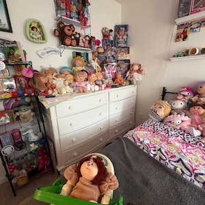 Könnte beinhalten: Ein Schlafzimmer mit einer großen Sammlung von Vintage-Puppen, darunter zahlreiche Cabbage Patch Kids, Plüschtiere und verschiedene Figuren, die auf einer weißen Kommode, einem schwarzen Metallregal und einem Bett mit gemusterter Decke stehen.