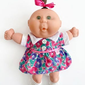 May include: A Cabbage Patch Kids doll with a round face, green eyes, and a small tuft of brown hair tied with a pink ribbon. The doll wears a white-collared dress featuring a floral pattern in pink, purple, and green, with two white buttons.