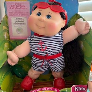 May include: A Cabbage Patch Kids doll with dark hair, blue eyes, and rosy cheeks, wearing a navy and white striped romper with a red anchor print, red shoes, and red sunglasses. The doll is in its original packaging with adoption papers.