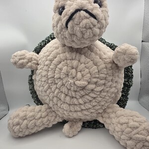 May include: A plush turtle toy crafted from chunky beige yarn with a textured, dark green and white speckled shell. The creature features a round head with black plastic eyes, a stitched black mouth, four limbs, and a small tail in the centre.
