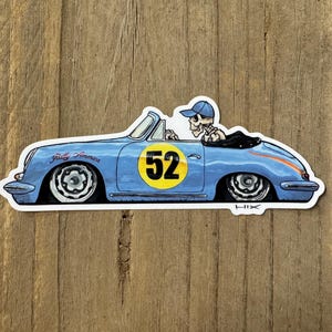 May include: A sticker of a light blue vintage convertible car with a skeleton driver wearing a blue cap. The car has a yellow circle with the number 52 and the words "Rally America". The sticker is on a wooden surface.