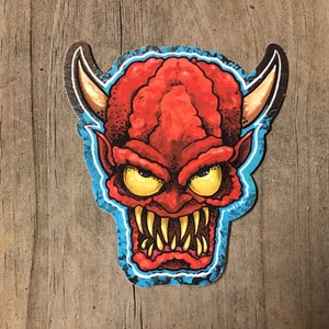 May include: Red and yellow cartoon devil face sticker with a blue outline. The sticker has a textured surface and is on a wooden background.