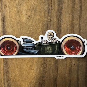 以下が含まれることがあります： 骸骨が運転する漫画のホットロッドのステッカー。車はオリーブグリーンで、赤いホイールとクロムエキゾーストパイプが付いています。「HTY Garage」の文字が車の側面に表示されています。