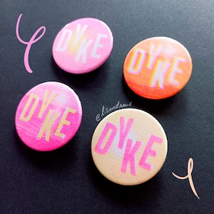 Peut inclure: Quatre badges ronds avec le mot "DYKE" en lettres roses et jaunes. Les badges présentent un dégradé de couleurs, notamment rose, orange et crème. Le fond est sombre.