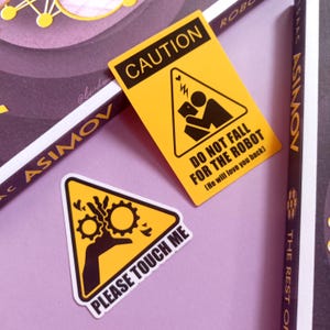 May include: Two yellow triangle stickers with black text and graphics. One says "CAUTION DO NOT FALL FOR THE ROBOT (He will love you back)". The other says "PLEASE TOUCH ME". The stickers are on a purple surface.