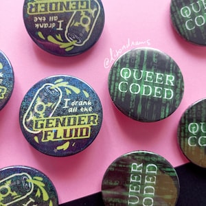 Può includere: Una collezione di spille rotonde con vari disegni. Alcune spille presentano il testo "I drank all the GENDER FLUID" con un'illustrazione di lattina. Altre spille mostrano "QUEER CODED" in un font in stile matrice. Le spille hanno uno sfondo scuro e iridescente.