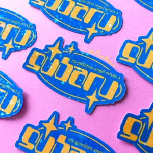 Könnte beinhalten: Blaue und gelbe Aufkleber mit dem Wort "Subaru" in Retro-Schrift, umgeben von Sternen und dem Satz "my guardian angel drives a." Die Aufkleber haben einen holografischen Effekt und liegen auf einem rosa Hintergrund.