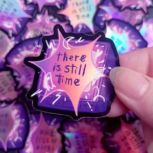 以下が含まれることがあります： 紫とオレンジ色のバーストデザインのホログラフィックステッカー。「there is still time」の文字が中央にあります。ステッカーは黒い縁取りと幽霊のイラストが特徴です。