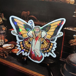 Puede incluir: Una pegatina holográfica que representa a una persona con alas de mariposa, sosteniendo dos cetros. La persona lleva un vestido blanco con detalles en rojo. Las alas son amarillas y negras con un diseño de telaraña.