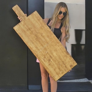 Puede incluir: Una tabla de cortar de madera rectangular grande con un asa. La tabla tiene un acabado marrón claro con vetas de madera visibles. El asa está unida a un extremo. La tabla mide aproximadamente 100 cm de largo y 30 cm de ancho.