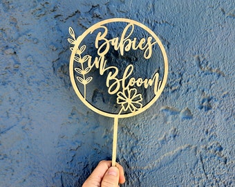 Puede incluir: Un círculo de madera con el texto "Babies in Bloom" y un diseño floral. El círculo está unido a un palo de madera.