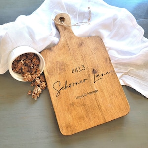 May include: A wooden cutting board with a handle, engraved with the text "4413 Schooner Lane Lloyd & Natalie".