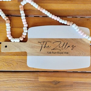 May include: A white and brown wooden serving board with a handle. The board is engraved with the text "The Zillas" and "728 Port Royal Ave".