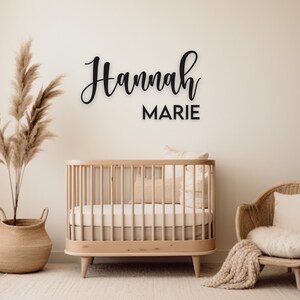May include: A nursery room with a wooden crib, a wicker chair with a white cushion, and a large dried pampas grass plant in a woven basket. The wall has a black wooden sign with the name "Hannah Marie" written in cursive.