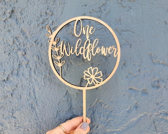 May include: Wooden cake topper with a circle design and the text "One Wildflower". The circle has a floral design and a small flower in the center.