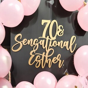 May include: A black background with gold lettering that reads "70s Sensational Esther." Pink balloons surround the sign. The sign is likely for a birthday celebration or party.