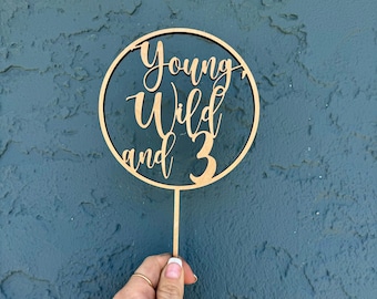 以下が含まれることがあります： 円形のデザインと「Young Wild and 3」の文字が書かれた木製ケーキトッパーです。