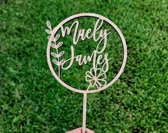 Puede incluir: Un adorno para pastel de madera con un diseño circular que presenta los nombres "Maely James" en escritura cursiva. El círculo está decorado con un diseño floral y una pequeña rama con hojas.