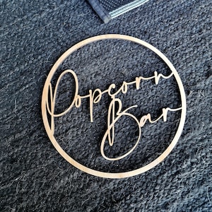 May include: A round wooden sign with the words "Popcorn Bar" in elegant cursive script. The sign is set within a circular frame. The sign is made of light-colored wood and is placed on a dark, textured surface.