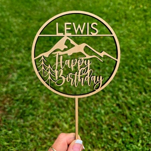 May include: A wooden birthday cake topper with the name "LEWIS" at the top. Below, a mountain range and trees are cut out, with "Happy Birthday" in cursive. The topper is held by a wooden stick.