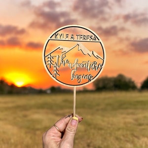 May include: Wooden cake topper with a mountain range silhouette and the text "Kyle & Teresa the adventure begins". The topper is on a stick and held by a hand against a sunset background.