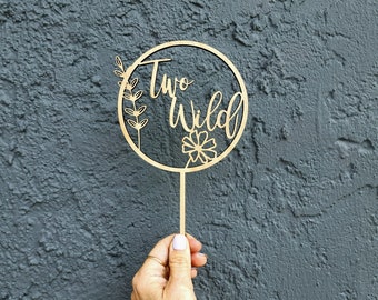 Puede incluir: Topper de pastel de madera con un diseño circular y las palabras "Two Wild" en una fuente cursiva. El círculo está decorado con una pequeña flor y hojas.