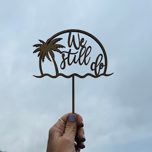 May include: Wooden cake topper with a palm tree and the words "We still do" in a cursive font. The topper is brown and has a white background.