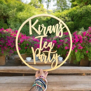 May include: A wooden circle sign with the text "Kiran's Tea Party" in gold lettering. The sign is held by a hand with multiple bracelets.