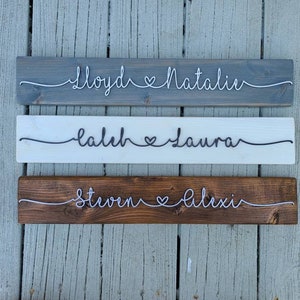 May include: Three wooden signs with names and a heart symbol written in white string. The top sign is gray, the middle sign is white, and the bottom sign is brown. The names on the signs are "Lloyd & Natalie", "Laleh & Laura", and "Steven & Alexi".