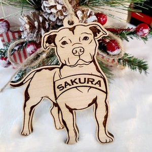 K&ouml;nnte beinhalten: Ein h&ouml;lzernes Hundeornament mit dem Namen "SAKURA" auf einem Banner eingraviert. Das Ornament ist hellbraun mit dunkleren braunen ge&auml;tzten Details und h&auml;ngt vor einer Weihnachtsdekoration mit Tannenzapfen und roten Beeren.
