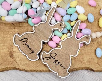 Op de afbeelding: Twee houten bunny-vormige ornamenten met de namen "Mommy" en "Jessie" erop geschreven. De ornamenten zijn omringd door pastelkleurige snoepjes in de vorm van paaseieren op een houten oppervlak.