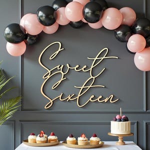 Puede incluir: Escena de fiesta con un letrero de madera "Sweet Sixteen", un arco de globos negros y rosas, cupcakes y un pastel peque&ntilde;o. El pastel est&aacute; decorado con bayas y una sola vela. El tel&oacute;n de fondo es una pared gris oscuro.