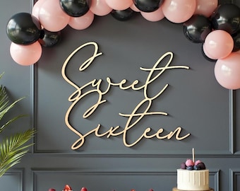 Puede incluir: Un letrero de madera "Sweet Sixteen" se muestra contra una pared gris oscuro, adornado con un arco de globos negros y rosas. Una mesa contiene cupcakes y un pastel pequeño con bayas.