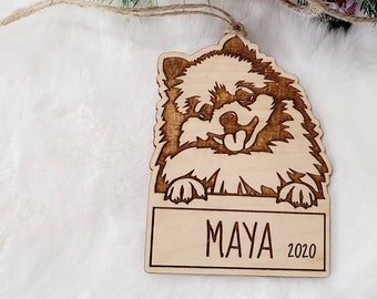 Könnte beinhalten: Ein Weihnachtsbaumschmuck aus Holz mit dem Gesicht eines Hundes, auf dem der Name "MAYA" und das Jahr "2020" auf einem Banner darunter eingraviert sind.