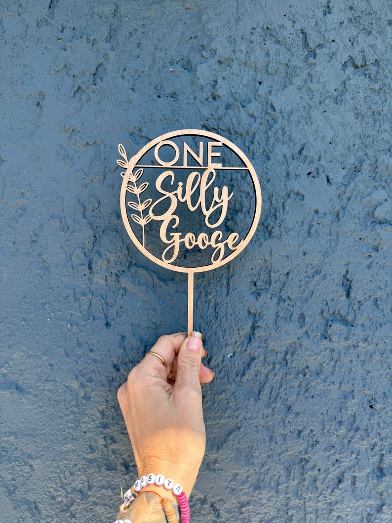 May include: A wooden cake topper with a circle design and the text "ONE Silly Gooze" in a cursive font. The circle has a leafy design on the left side.
