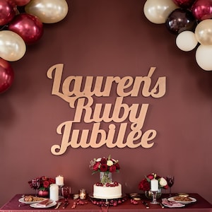 Op de afbeelding: Een feestelijke scène met een bordeauxrode achtergrond, versierd met bordeauxrode, crème en bruine ballonnen. De woorden "Lauren's Ruby Jubilee" worden in een cursief lettertype weergegeven. Een tafel is gedekt met een taart, bloemen, kaarsen en borden.