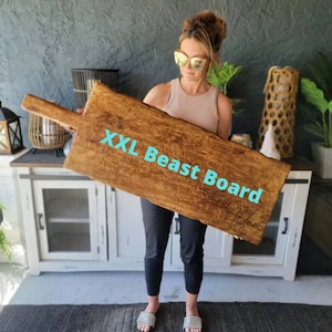Puede incluir: Una tabla de servir de madera rectangular grande con un asa. La tabla es de color marr&oacute;n c&aacute;lido con vetas de madera visibles y el texto "XXL Beast Board" en turquesa. La tabla es sostenida por una persona.