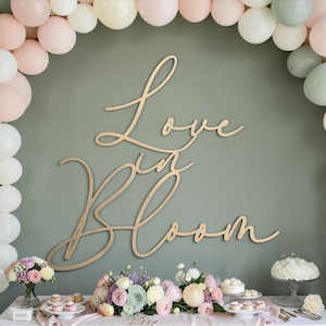 May include: A wooden sign with the words "Love in Bloom" in cursive script is mounted on a sage green wall. The sign is surrounded by pastel balloons and a table with floral arrangements, cupcakes, and a cake.