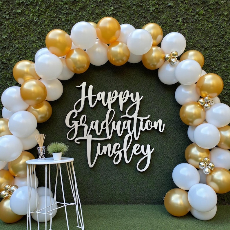 Puede incluir: Un tel&oacute;n de fondo para una fiesta de graduaci&oacute;n con un arco de globos dorados y blancos. Un letrero blanco con las palabras "Happy Graduation Tinsley" est&aacute; centrado. Una peque&ntilde;a mesa blanca con una planta y decoraci&oacute;n est&aacute; al frente.