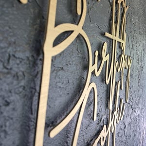 Puede incluir: Un letrero de madera con las palabras "Happy Birthday Sophie" en escritura cursiva. El letrero est&aacute; hecho de madera de color claro y est&aacute; montado sobre una superficie texturizada de color gris oscuro. Las letras est&aacute;n conectadas.