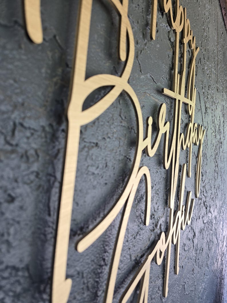Puede incluir: Un letrero de madera con las palabras "Happy Birthday Sophie" en escritura cursiva. El letrero est&aacute; hecho de madera de color claro y est&aacute; montado sobre una superficie texturizada de color gris oscuro. Las letras est&aacute;n conectadas.